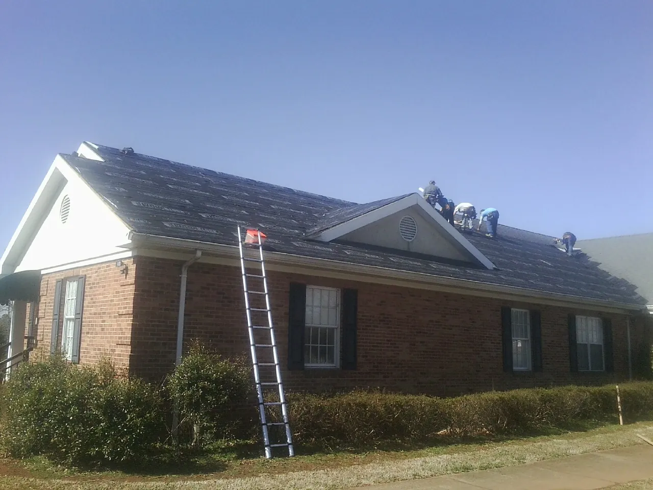 Crew tearing shingles off a brick ranch roof with a ladder set — Emergency Roof Repair work by our crew in Bat Cave