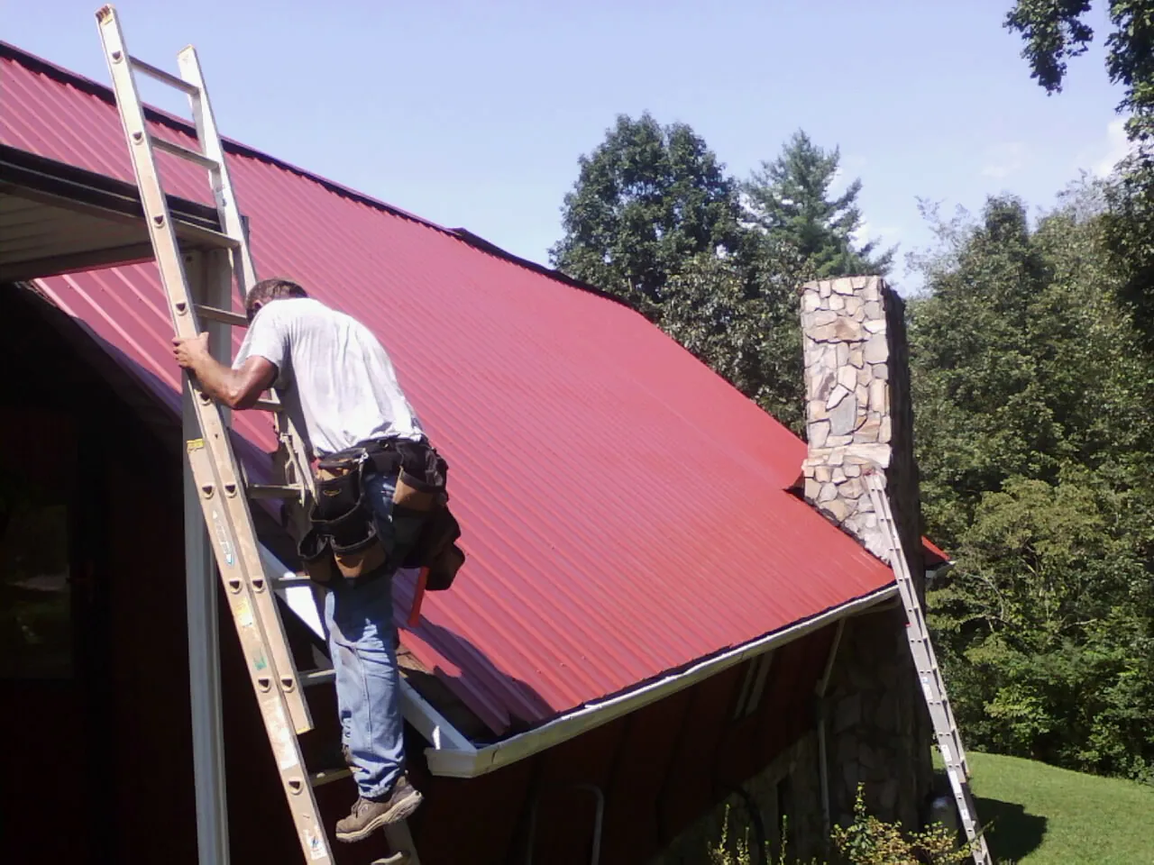 Roofer on a ladder beside a red metal roof and stone chimney — Soffit & Fascia Repair by Roofing Brothers in Bat Cave